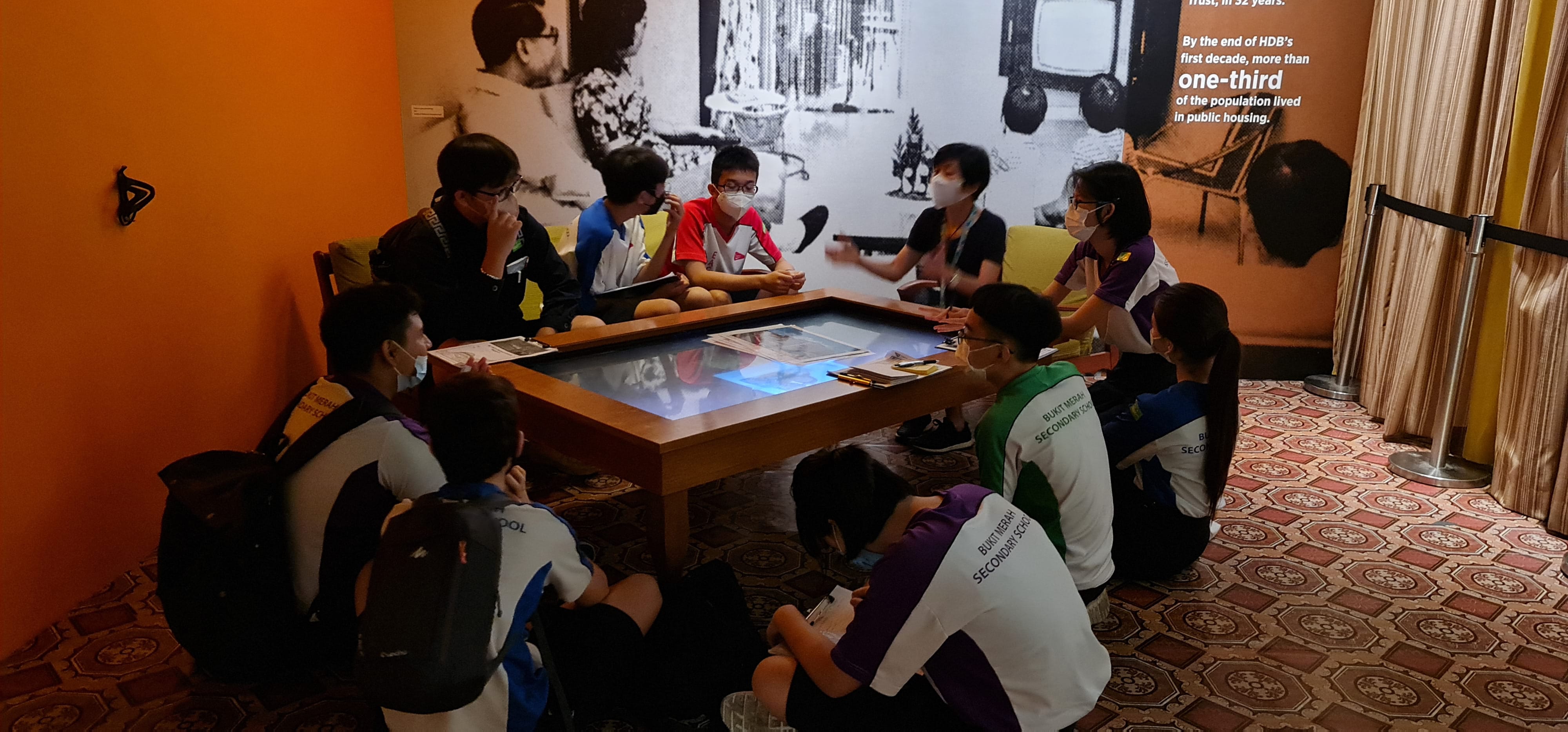 Students in school uniforms gather around a lit interactive table in a museum exhibit about HDB housing.